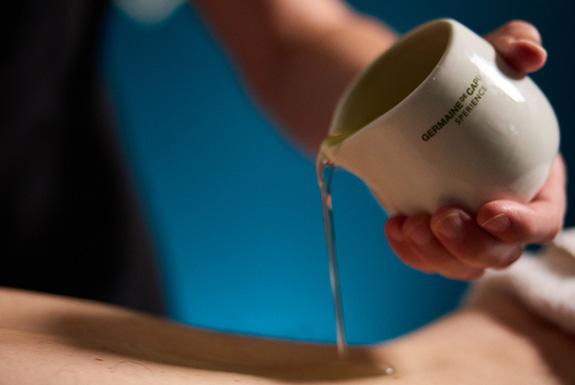 Person pouring oil from pouring jug onto a person&#039;s back.